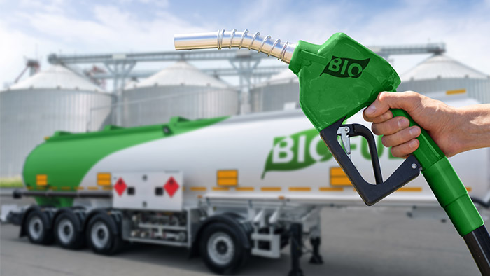 Hand holding a green biofuel pump nozzle in front of a biodiesel tanker and storage tanks